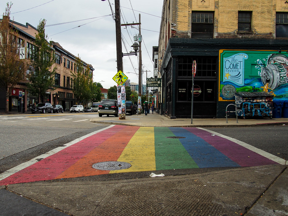 A view of a rainbow-painted crosswalk in the Capitol Hill neighborhood of Seattle. There is litter in the street and on the crosswalk.