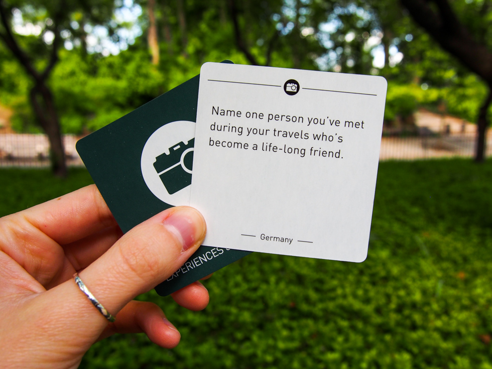 Hand holding Paths Crossing Experiences & Encounters card that reads "Name one person you've met during your travels who's become a life-long friend."