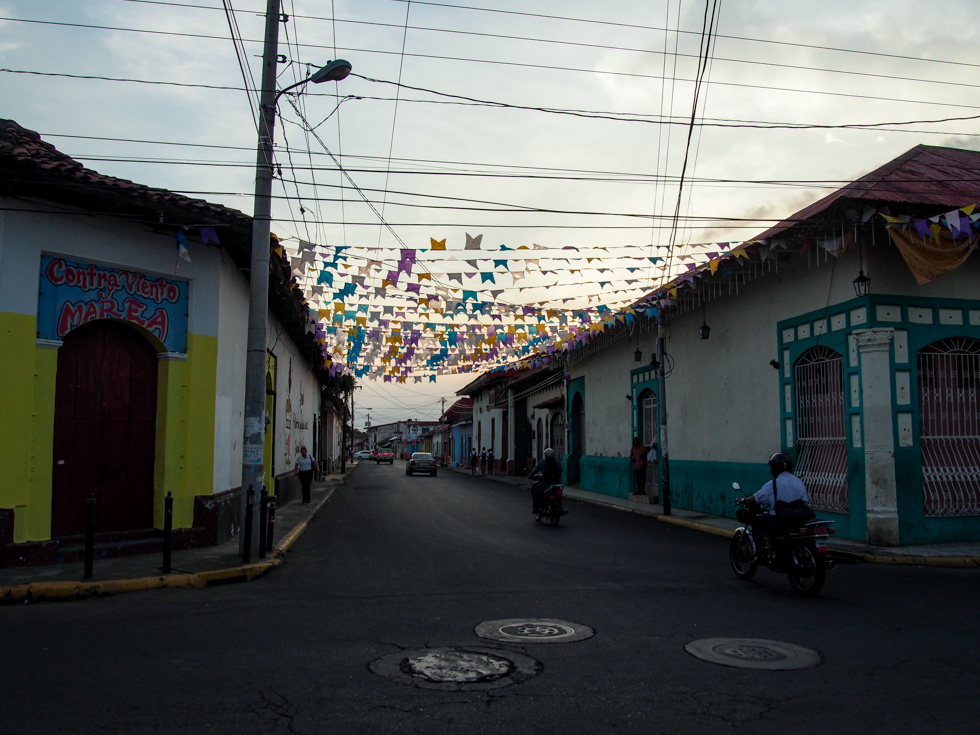 Streets in Leon, Nicaragua at dusk, with some colorful flags overhead flapping in the breeze and motorcyclists passing on the street