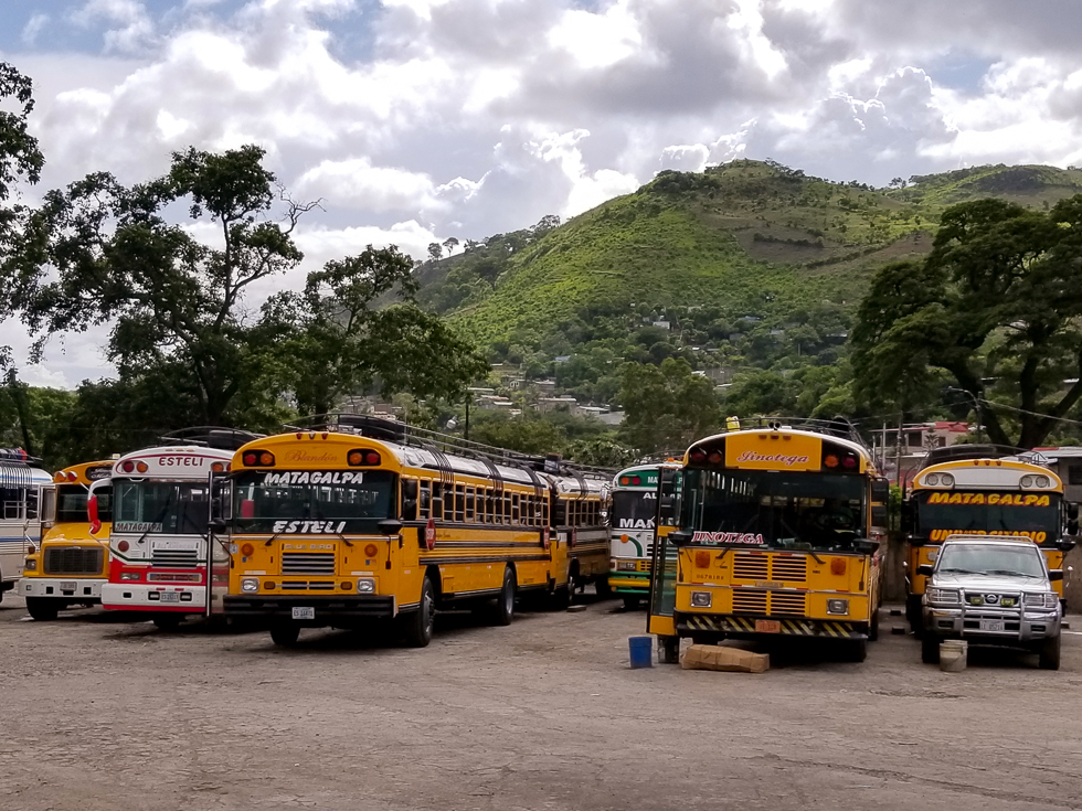 public transportation in Nicaragua: a row of chicken buses (retrofitted yellow school buses) in Matagalpa's bus station, with a mountainous background and a blue, cloudy sky