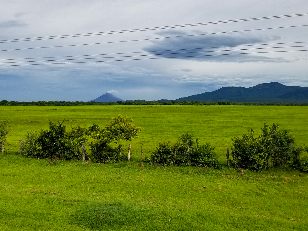 View of a lush green field with a smoking volcano in the distance, seen from a public bus in Nicaragua