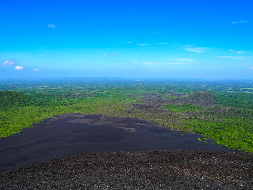 View of the Nicaraguan landscape from the top of the cerro negro volcano - green hills, a sliver of the city of Leon in the background, and the Pacific ocean in the distance, under a clear blue sky
