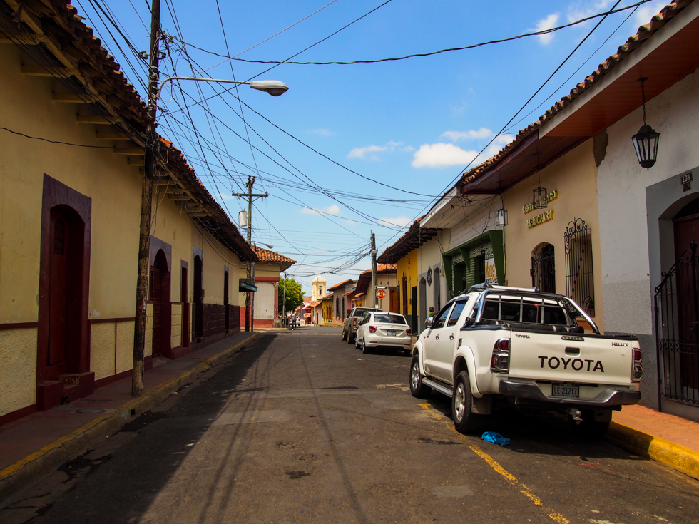 View of a quiet street in Leon, Nicaragua, with rows of tan, colonial buildings and a few parked cars, under a sunny, blue sky