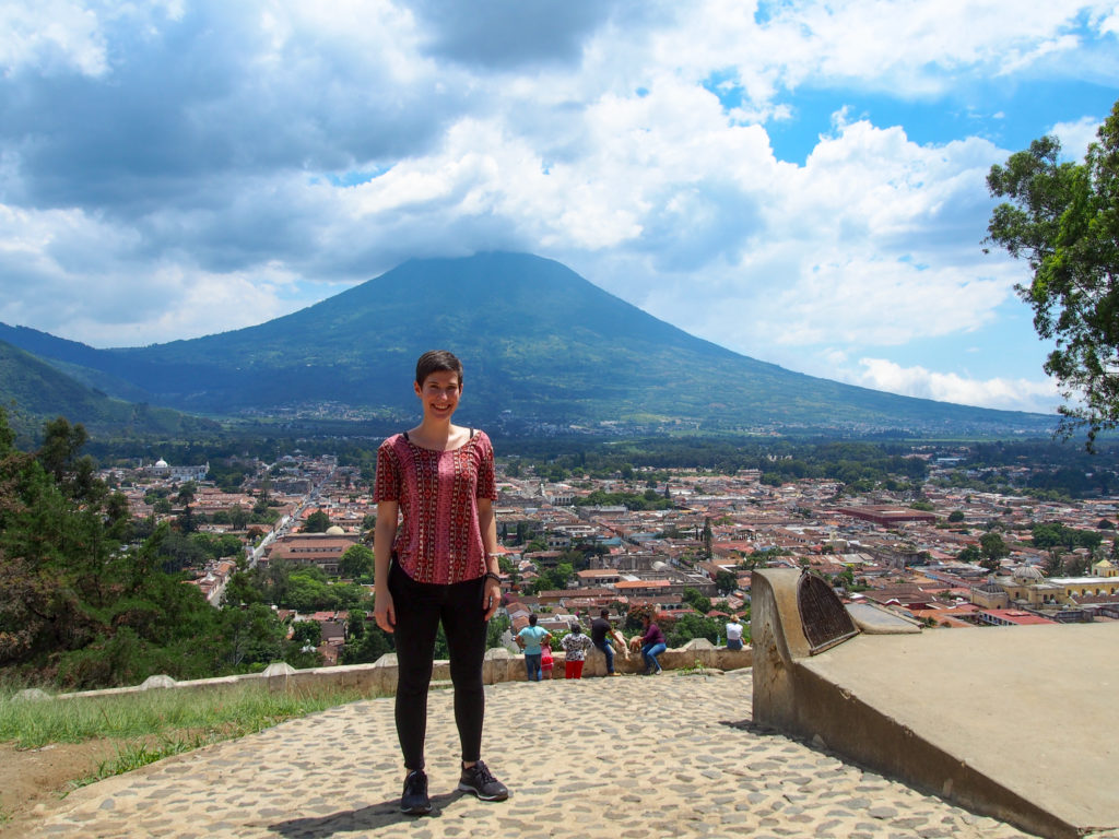 Alissa smiling in the sun at the cerro de la cruz - she is wearing a pink shirt and black pants, and behind her is the city of Antigua and the Water Volcano