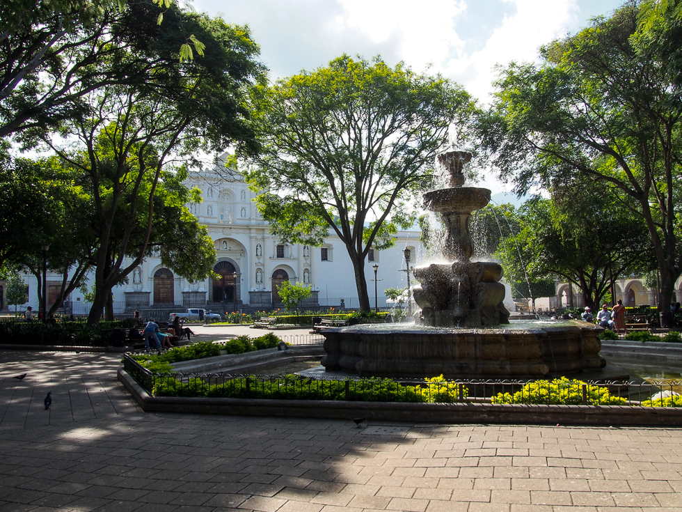 Antigua, Guatemala's central parque in the morning: view of the fountain in the center, some green trees, and the cathedral in the background