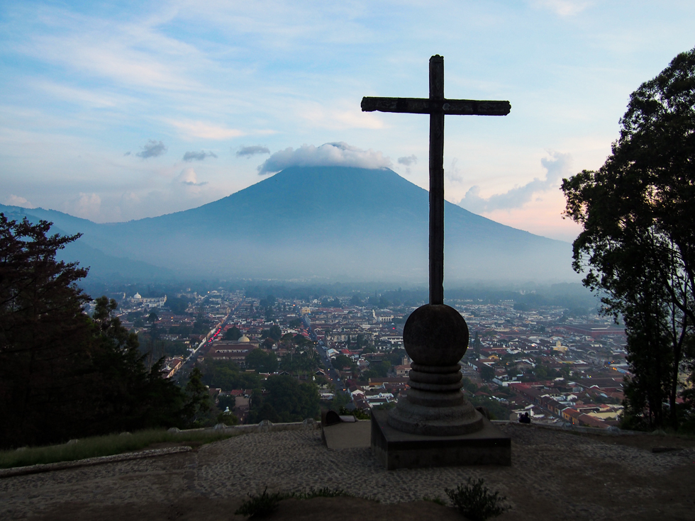 View from Antigua, Guatemala's cerro de la cruz at sunset: a large cross in the foreground, in the distance the city of Antigua, followed by the Water Volcano in the background